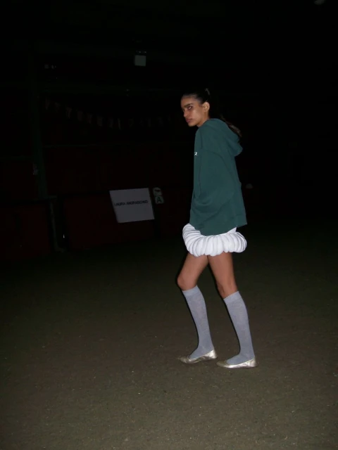 Young woman wearing a green hoodie, white skirt, and gray socks stands outside in a dark environment looking at the camera.