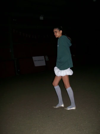 Young woman wearing a green hoodie, white skirt, and gray socks stands outside in a dark environment looking at the camera.