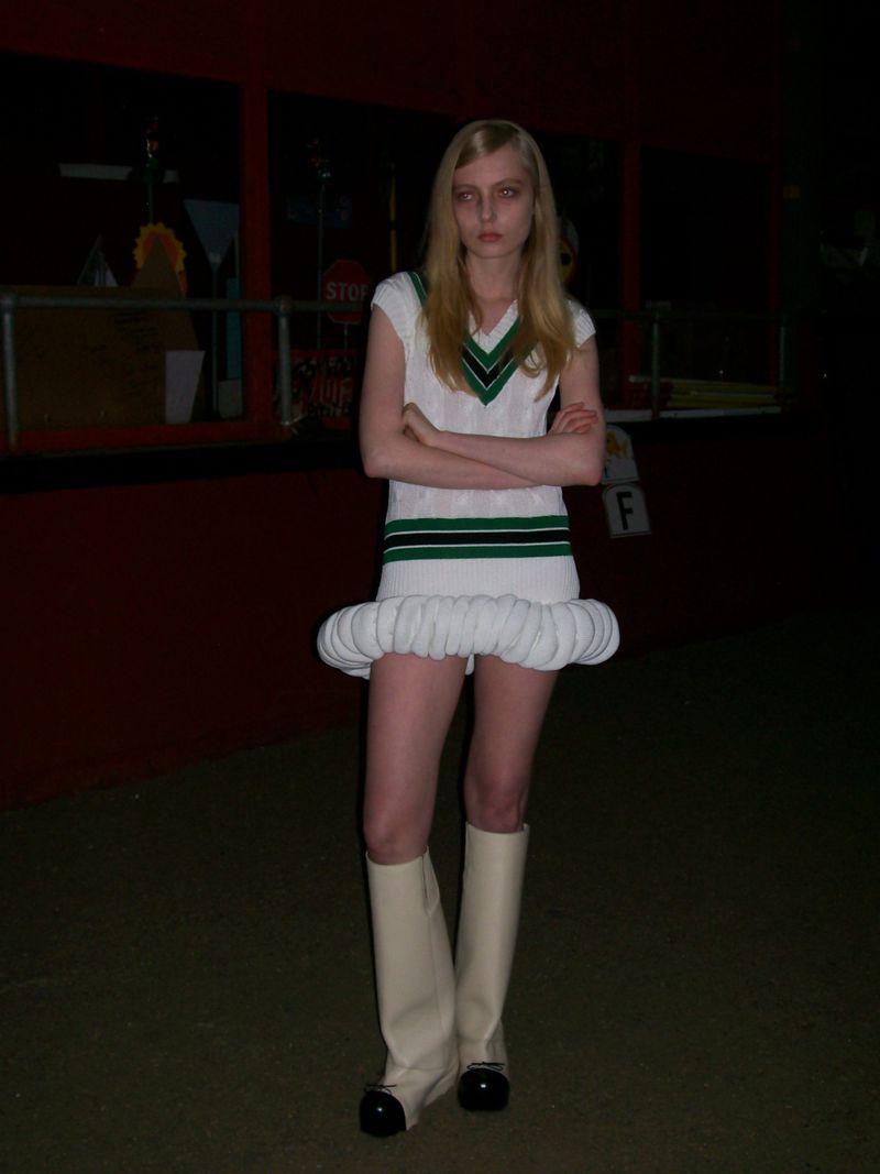 Young woman in a white V-neck sweater and short dress with puffed hemline, wearing white boots, stands with her arms crossed.