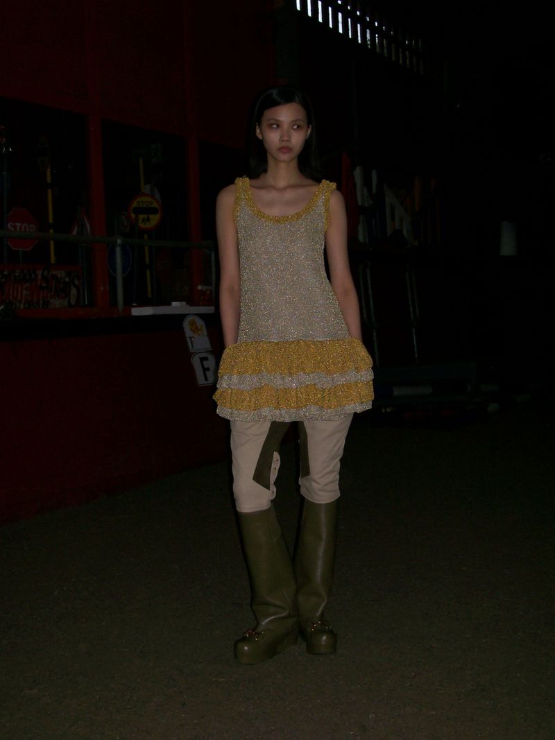 Woman stands in a textured gold and yellow ruffled dress with khaki pants and olive green platform boots in an urban setting at night.