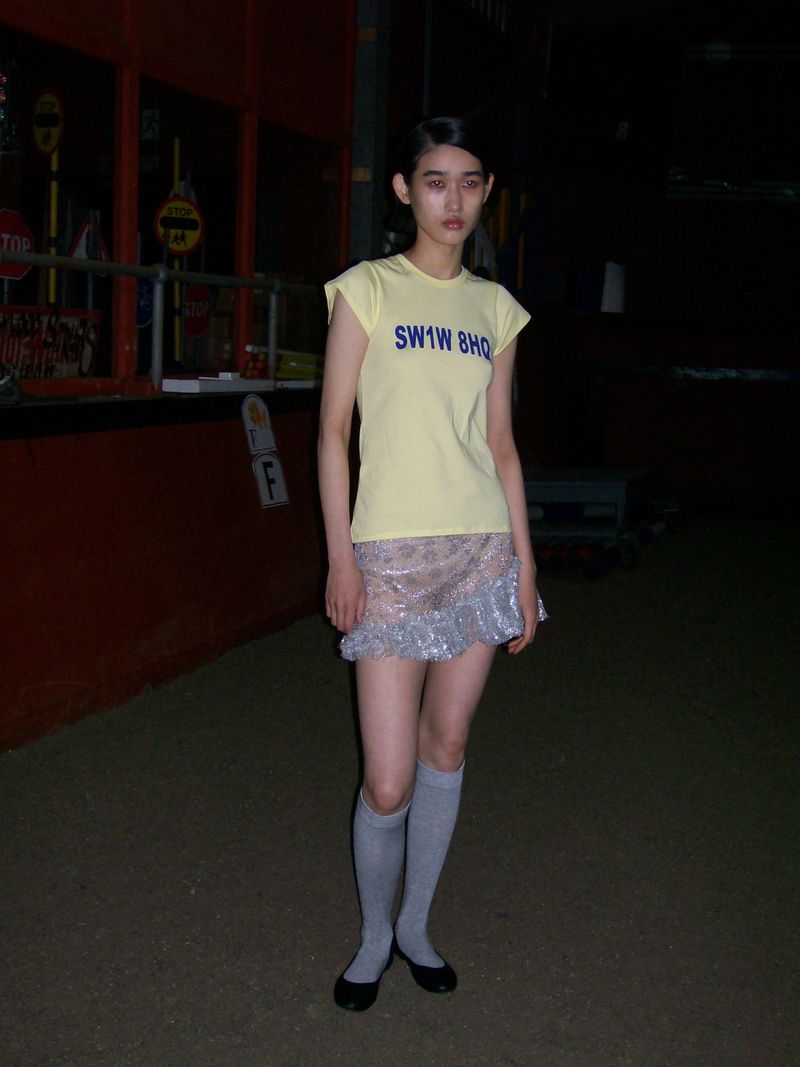 Young woman in a yellow top and silver skirt stands in an outdoor setting with dark lighting, creating a unique fashion portrait.
