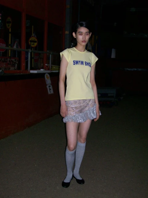 Young woman in a yellow top and silver skirt stands in an outdoor setting with dark lighting, creating a unique fashion portrait.