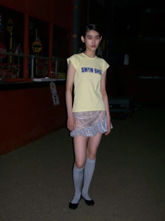 Young woman in a yellow top and silver skirt stands in an outdoor setting with dark lighting, creating a unique fashion portrait.