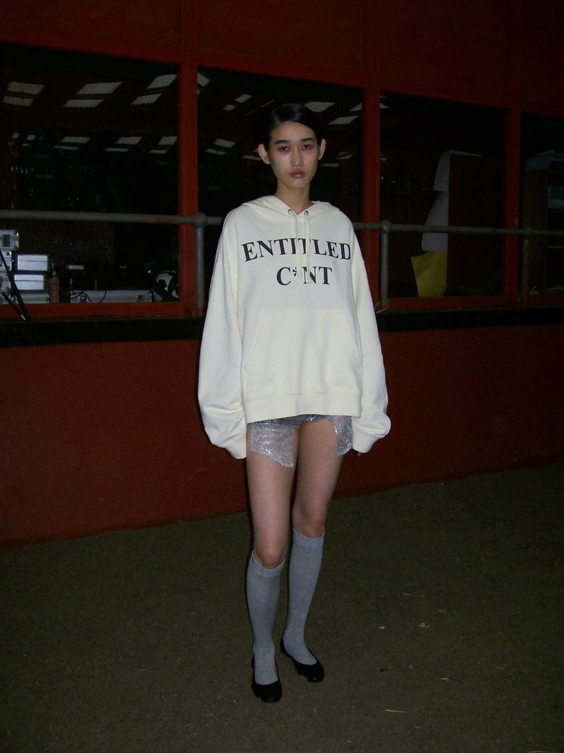 A solitary young woman in a hooded sweatshirt, mini skirt, knee socks and flats poses against a dimly lit, red and black background.
