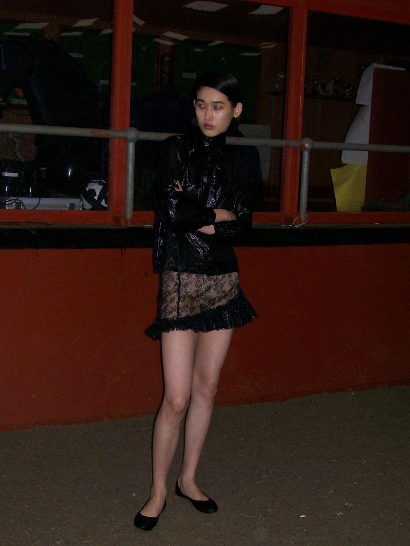 Young woman in black jacket and lace skirt poses against a red wall in an eye-level medium shot. Fashion editorial.