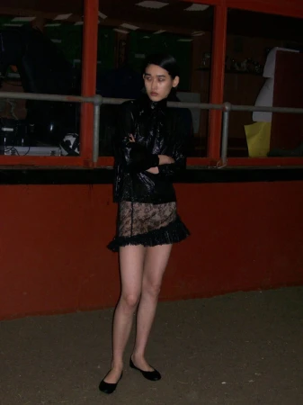 Young woman in black jacket and lace skirt poses against a red wall in an eye-level medium shot. Fashion editorial.