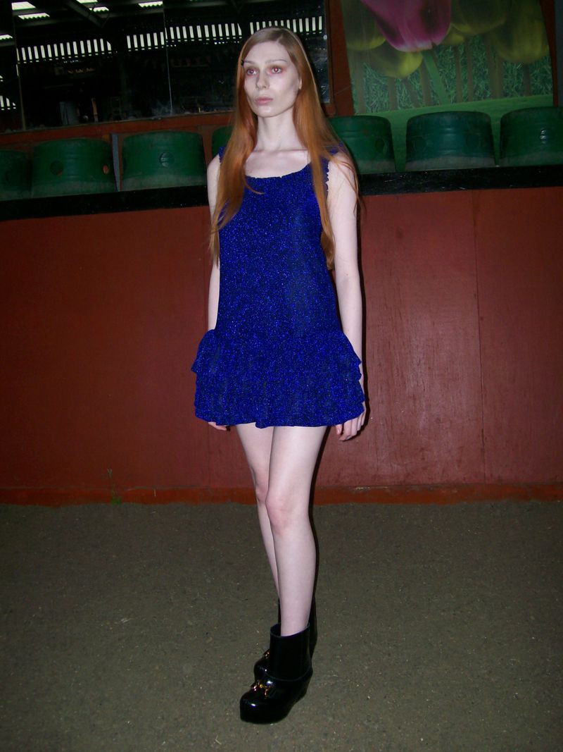 A slender young woman with long red hair poses in a sparkly blue minidress and black platform boots against a muted indoor backdrop.
