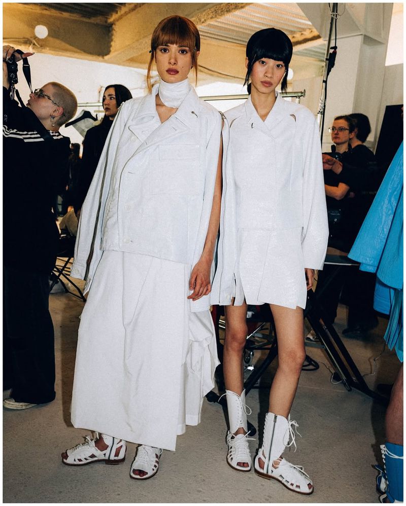 Two fashion models in white outfits pose in a studio setting. They are wearing matching open-toe shoes and various garments.