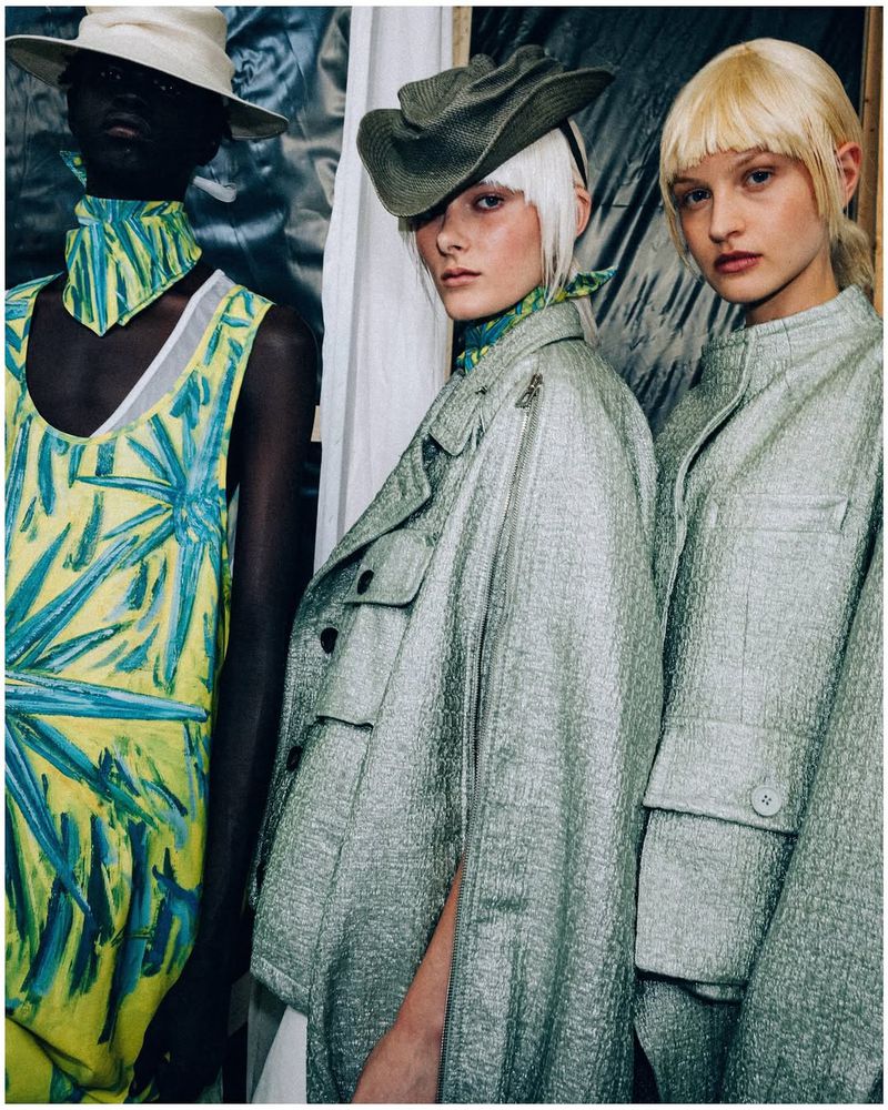 Three models backstage at the SONGZIO S/S 25 show wearing textured sage green outerwear and a vibrant painted yellow tunic.