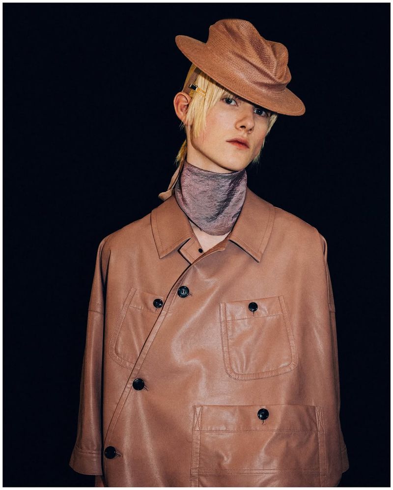 Androgynous person wearing a light brown hat and jacket against a black background in a studio shot. Fashion concept.