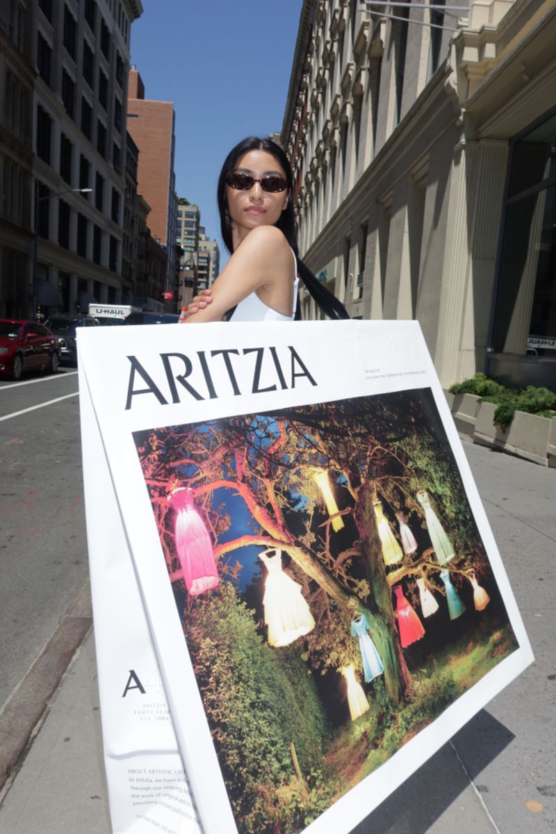 Woman poses with Aritzia advertisement in a city street with tall buildings and vehicular traffic under a clear blue sky.