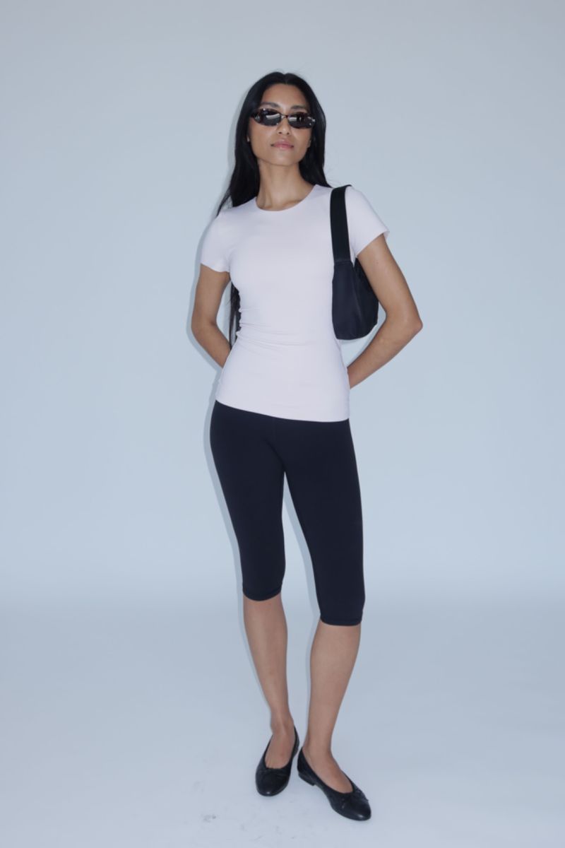 A full shot of a woman with sunglasses, a pink t-shirt, black leggings, and ballet flats, posing against a white backdrop.