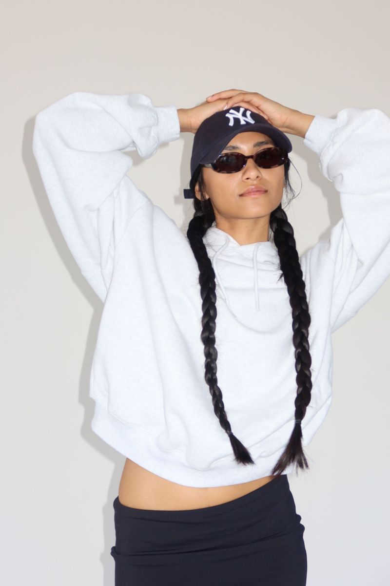 A woman with braided hair wearing sunglasses and a baseball cap poses in a white sweatshirt and a dark skirt against a plain background.