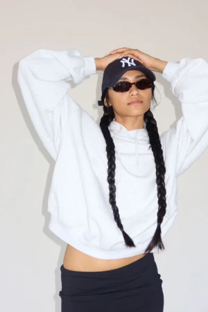 A woman with braided hair wearing sunglasses and a baseball cap poses in a white sweatshirt and a dark skirt against a plain background.