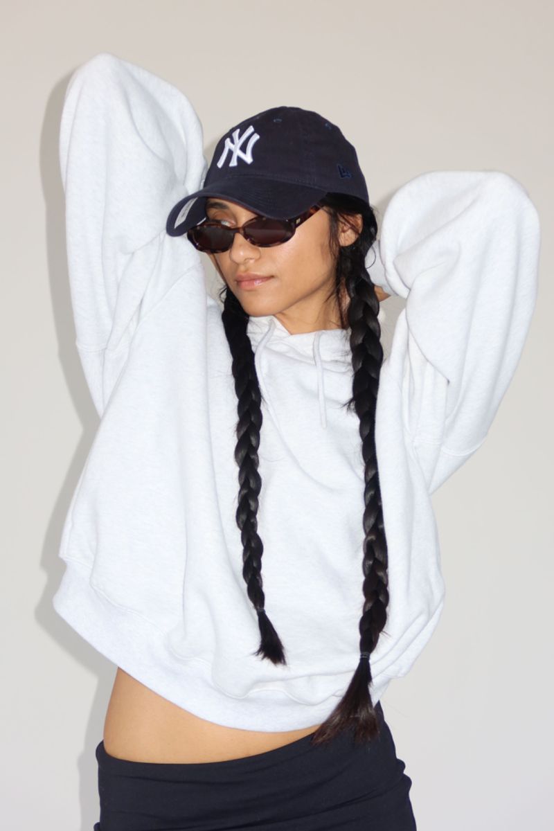 Young adult woman in a white hoodie and New York Yankees cap poses in front of a neutral wall, with long dark braids and sunglasses.