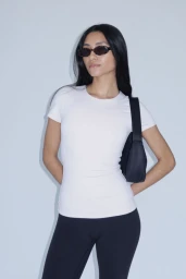 Model Alvina Bokhari wears a white fitted t-shirt, dark sunglasses, and a black shoulder bag against a neutral studio backdrop.