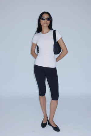 Model Alvina Bokhari in a white fitted t-shirt, black capri leggings, sunglasses, and black flats against a white studio background.