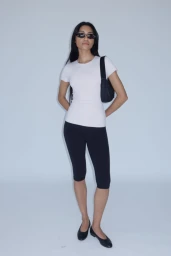 Model Alvina Bokhari in a white fitted t-shirt, black capri leggings, sunglasses, and black flats against a white studio background.