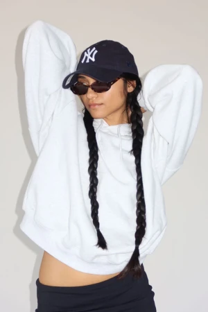 Alvina Bokhari wearing an oversized white hoodie, dark sunglasses, and a navy baseball cap with twin braids against a studio background.