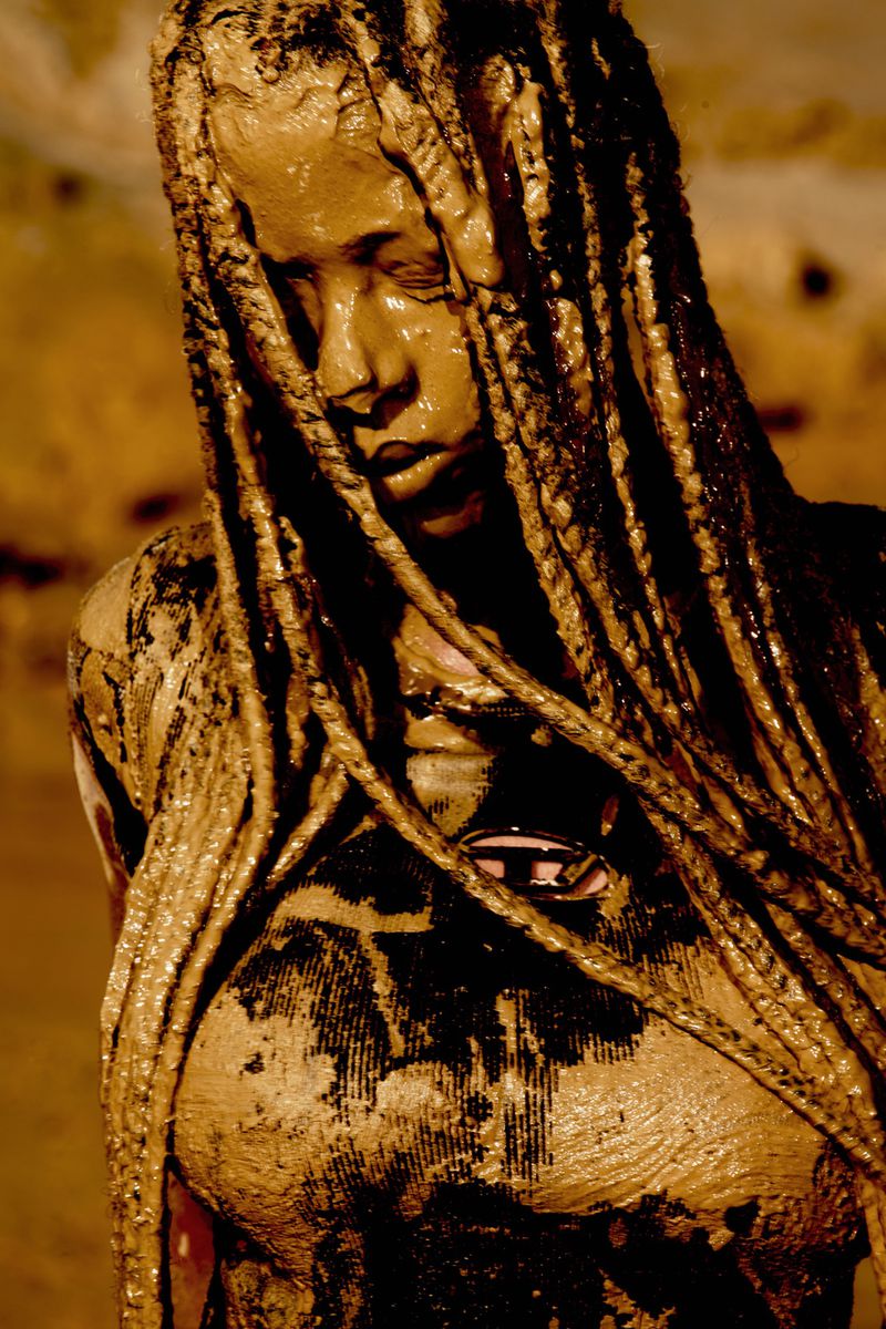 Model completely covered in thick wet brown mud with dreadlocks framing the face and a metallic logo badge on the chest.