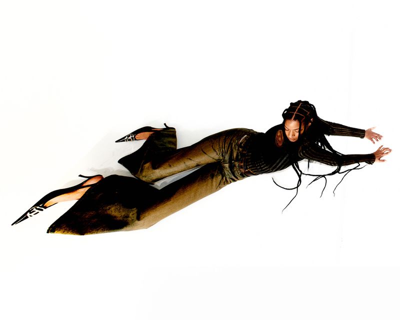 A woman lies against a white background wearing a long-sleeved black top, brown pants, and zebra-print high heels, extending her arms and legs.
