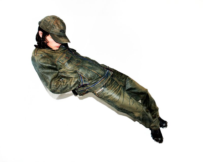 Person in worn denim jacket and pants, lying on a white surface, looking away from the camera in a fashion-style pose. 