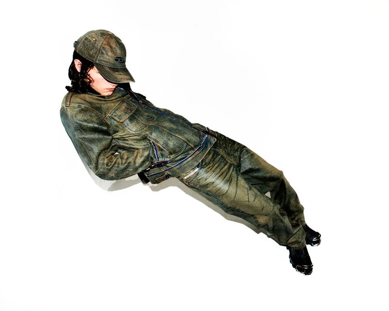 Person in worn denim jacket and pants, lying on a white surface, looking away from the camera in a fashion-style pose. 