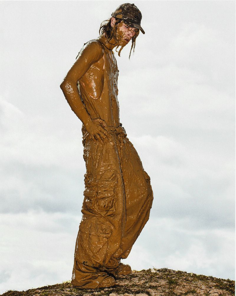 Adult male wearing camouflage cap and brown overalls covered in mud posing outdoors in front of a cloudy, overcast sky.
