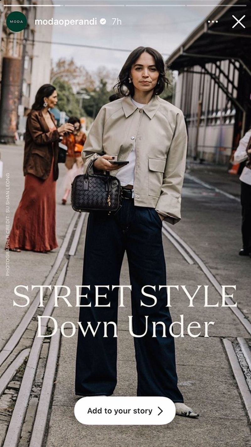 A fashionable woman in casual chic style stands on a city street with blurred figures, train tracks, and industrial architecture in the backdrop.
