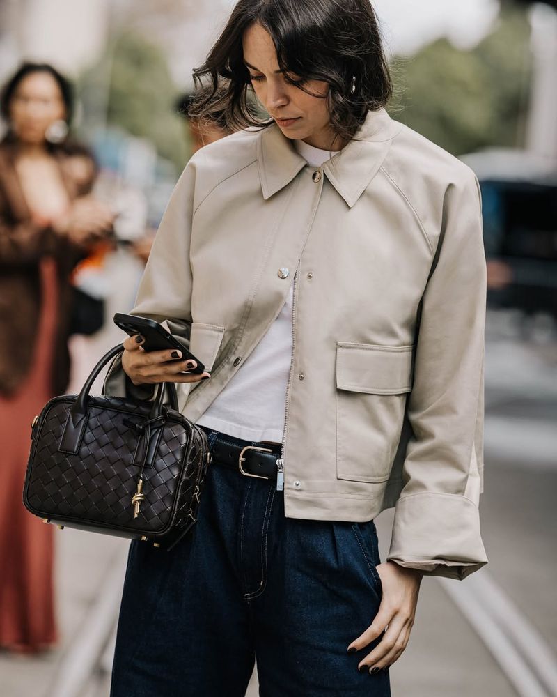 Brunette woman in a beige jacket and blue jeans holds a smartphone and purse, with a blurred background of a city street.