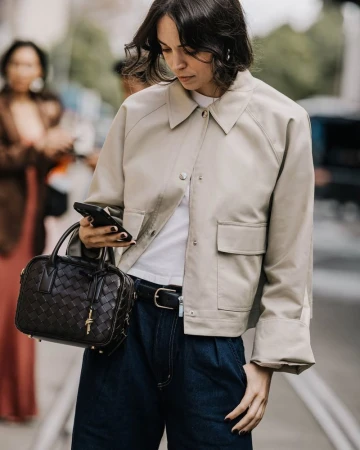 Woman in a beige cropped jacket holding a woven black handbag and looking down at a smartphone on a city street.