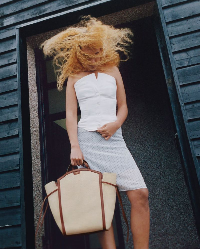 Blonde woman in white top and skirt stands in a doorway holding a tan tote bag, with her hair windswept and blowing around her face.