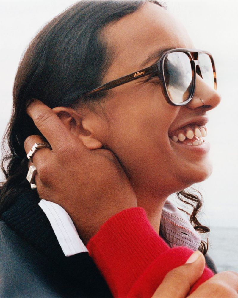 A young adult smiles outdoors in a medium shot, wearing aviator sunglasses and silver rings with a hand gently touching her face.