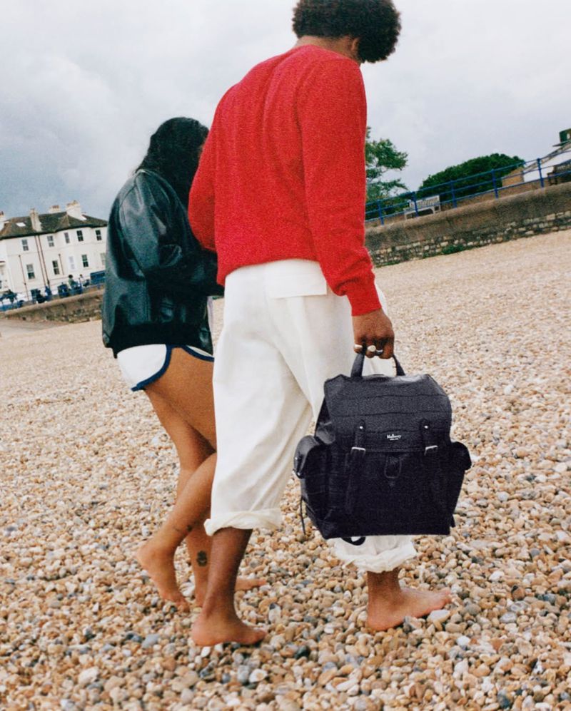 Couple walking barefoot on a pebble beach with a backpack. He is wearing a red shirt and white pants; she is in a jacket and shorts.