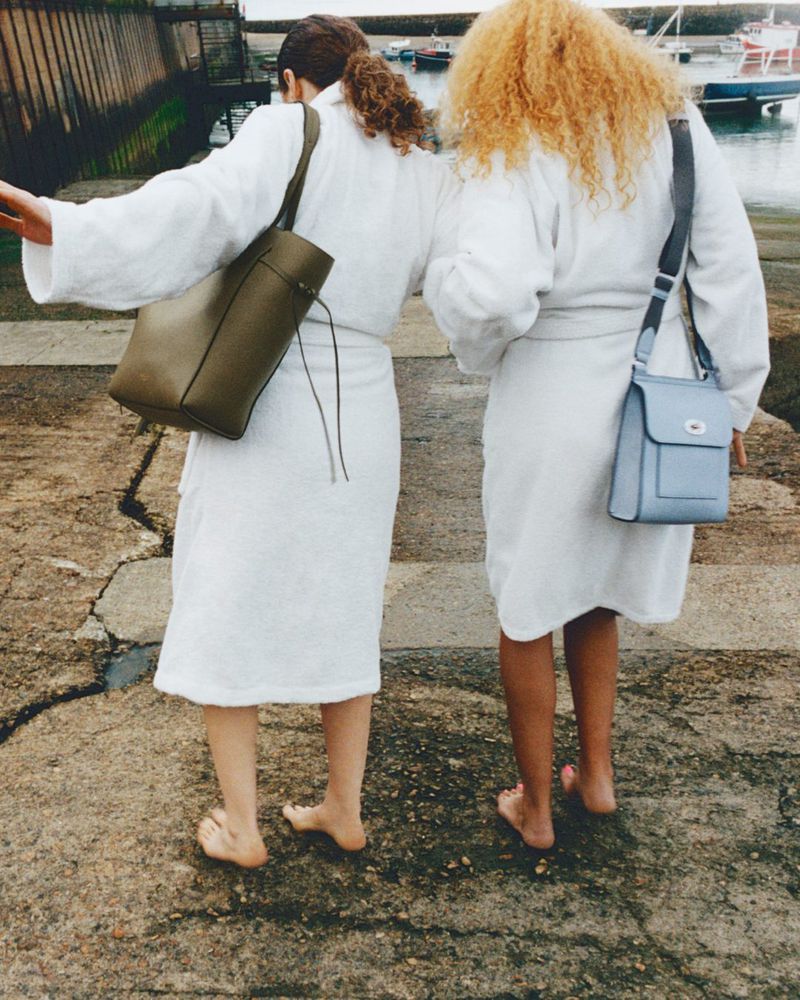 Two barefoot women in white robes walk on a stone pier by the sea, carrying shoulder bags, with boats and a harbor in the background.