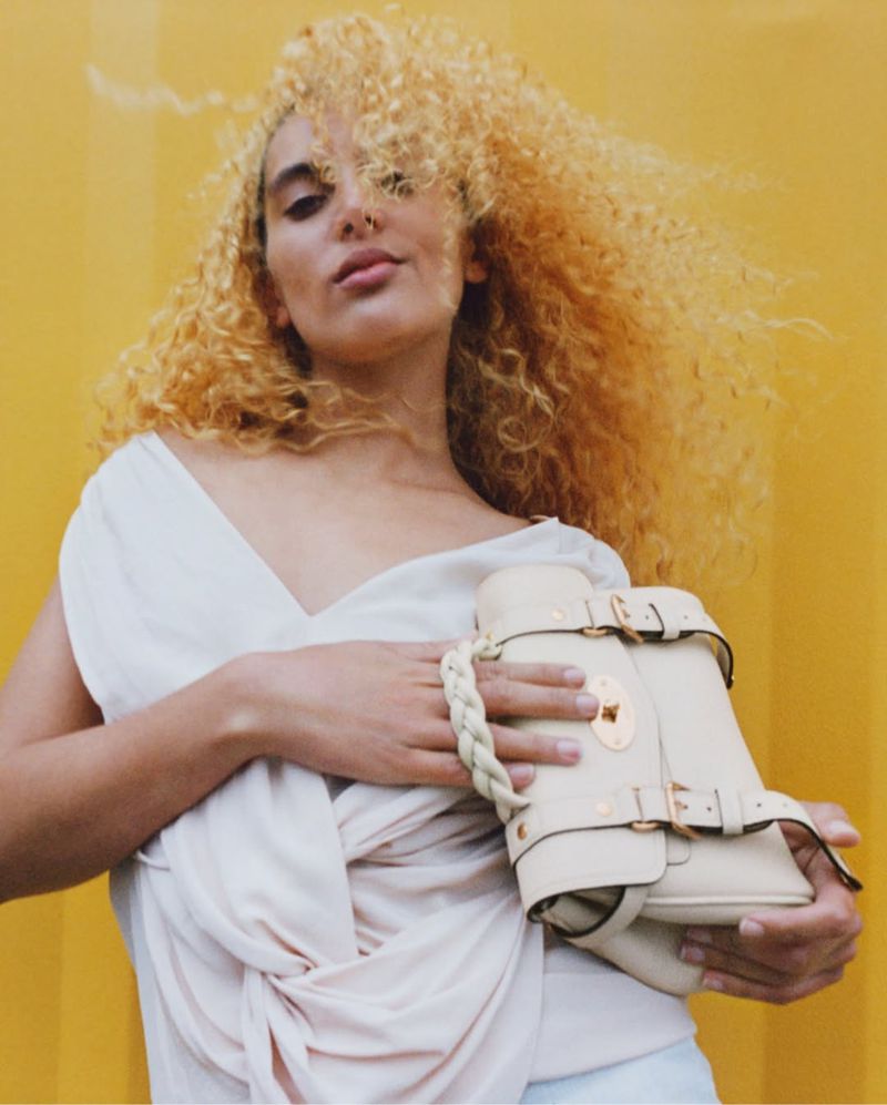 A woman with curly hair poses with a beige leather handbag against a yellow background in a fashion or beauty style portrait shot.