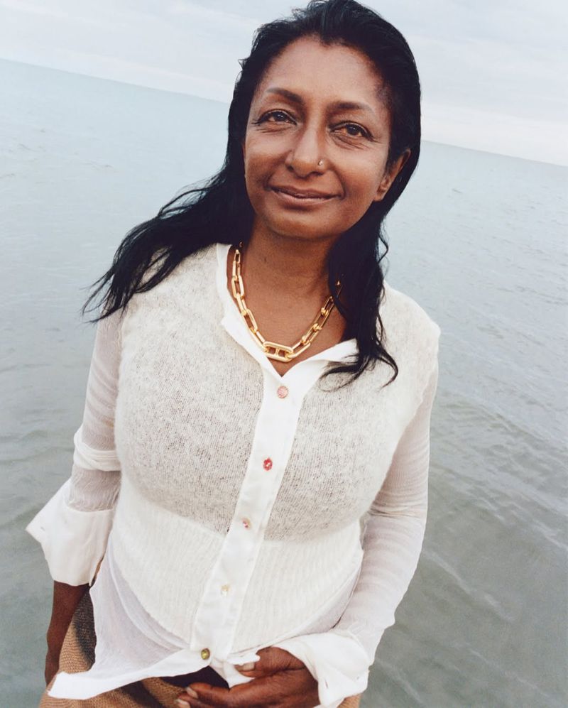 Portrait of an adult woman with dark hair and a gold necklace, standing on a beach near the water with an overcast sky in the background.