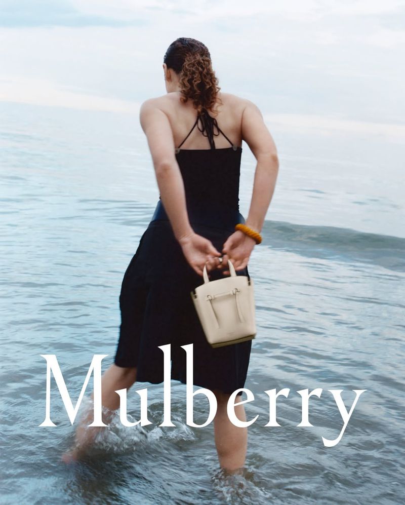 A woman in a black dress stands in the ocean, holding a cream-colored handbag with the text 'Mulberry' overlayed on the image.