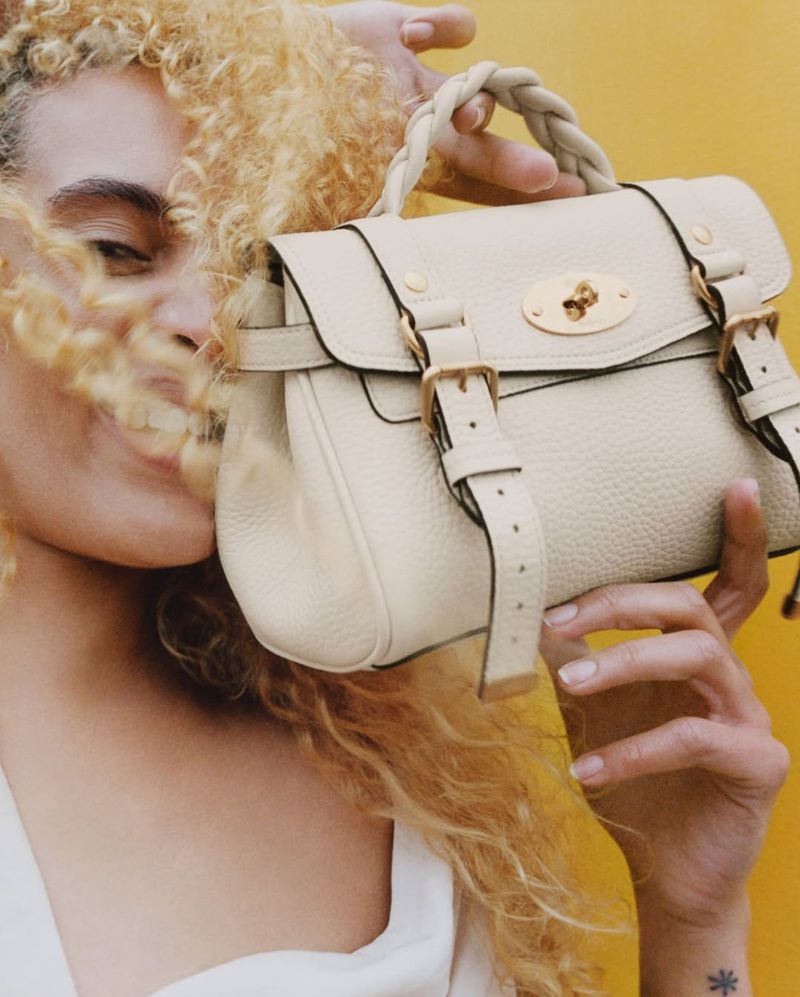 Close-up of an adult woman with blonde curly hair holding a cream-colored leather handbag with gold hardware against a yellow background.