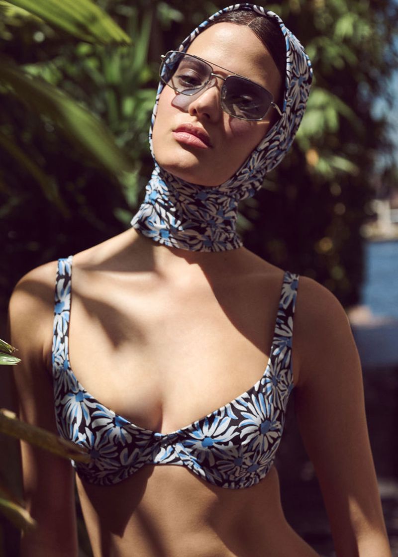 Woman in floral bikini and headscarf poses in a tropical setting with bright sunlight casting shadows through lush green foliage.