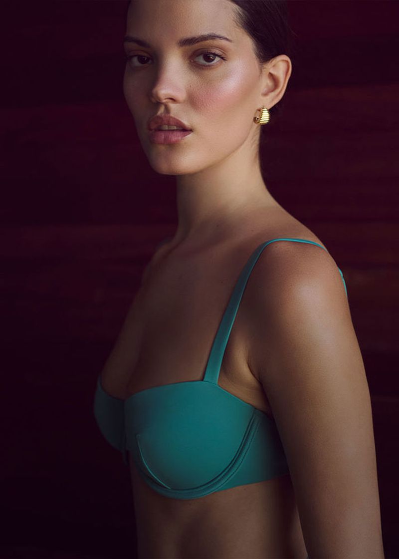 Close-up of an adult woman wearing a teal bra and gold earrings, posing in a dark studio. Beauty and fashion photography.