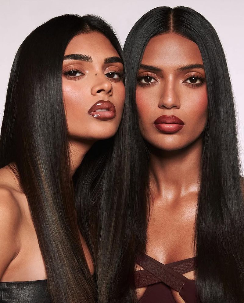 Two women with long, straight dark hair pose indoors against a light background in a beauty shot, showcasing glossy lips and makeup.
