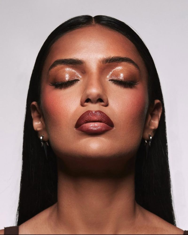 Close-up beauty shot of an adult female model with dark hair and brown skin, wearing shiny brown lipstick and shimmery makeup.