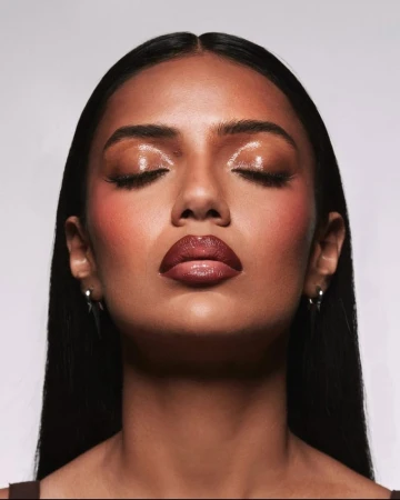Close-up beauty shot of an adult female model with dark hair and brown skin, wearing shiny brown lipstick and shimmery makeup.