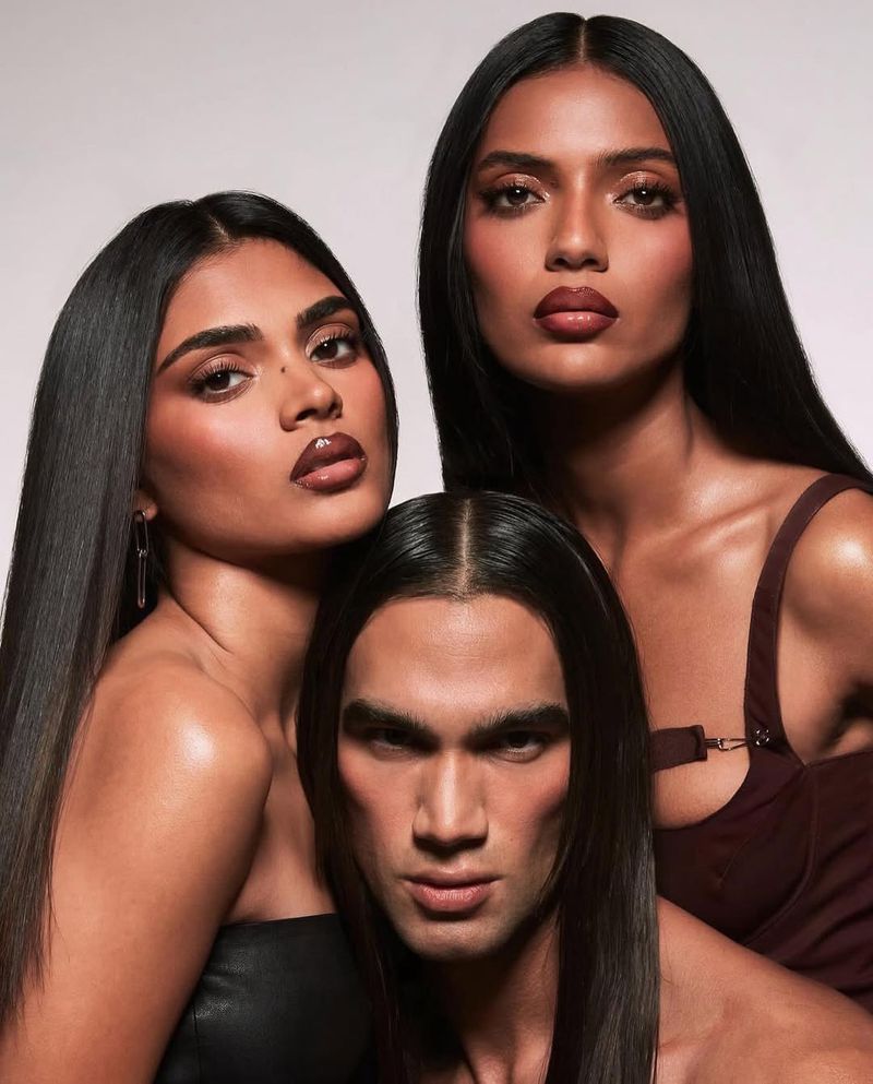 Three models pose in a studio. Two women and one man with straight dark hair. The women wear brown lipstick and strapless tops.