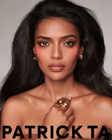 Close-up portrait of a woman with brown hair wearing gold jewelry, featuring pink blush and nude lips, against a neutral background.
