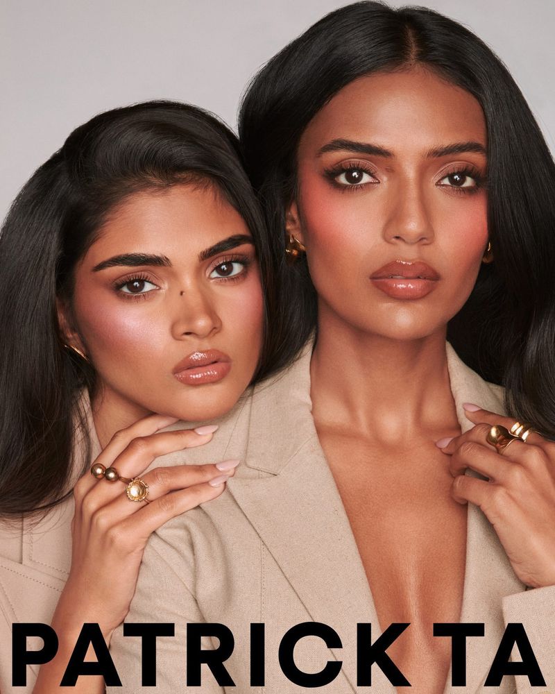 Two dark-haired women pose closely in beige jackets. Makeup is prominent, with glossy lips, flushed cheeks. Rings adorn their manicured hands.