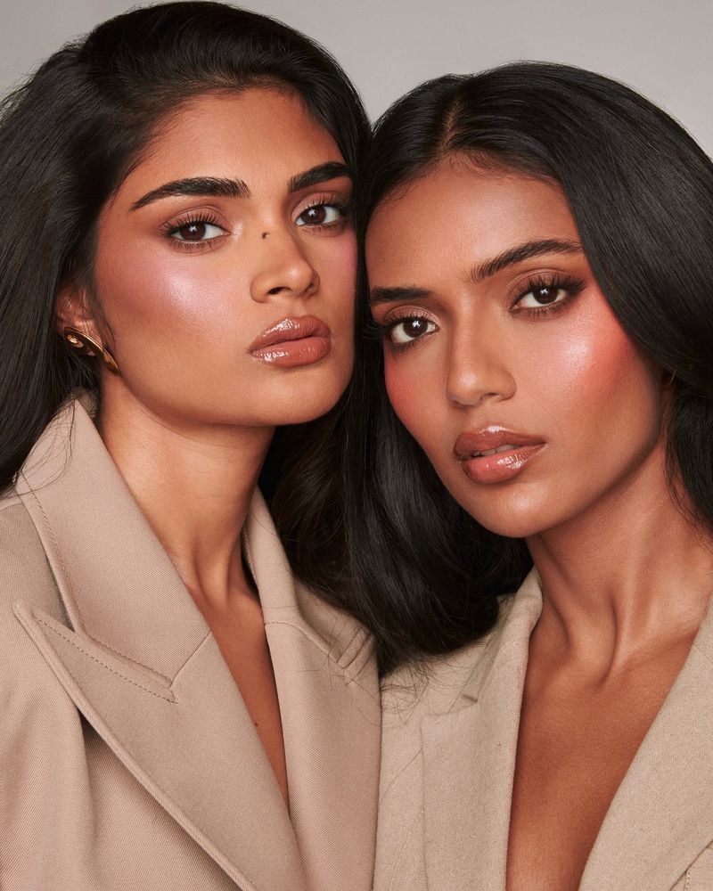 Close-up studio shot of two beautiful South Asian women with dark hair and tan blazers, showcasing elegant makeup in a bright, neutral setting.