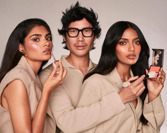 Three people pose indoors in a beauty-style photo: two dark-haired women with makeup, and a man in the center with glasses, all in beige blazers.