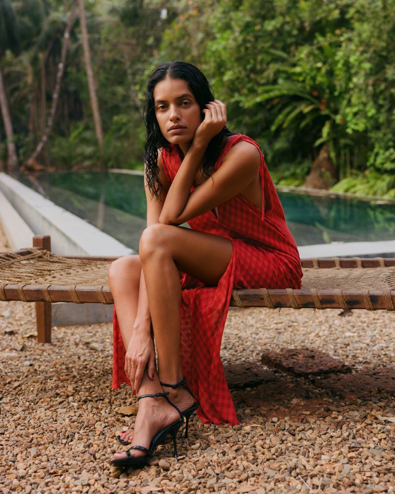 Adult woman in a red dress sits on a wooden bench with black strappy high heels and wet hair in an outdoor tropical setting by a pool.